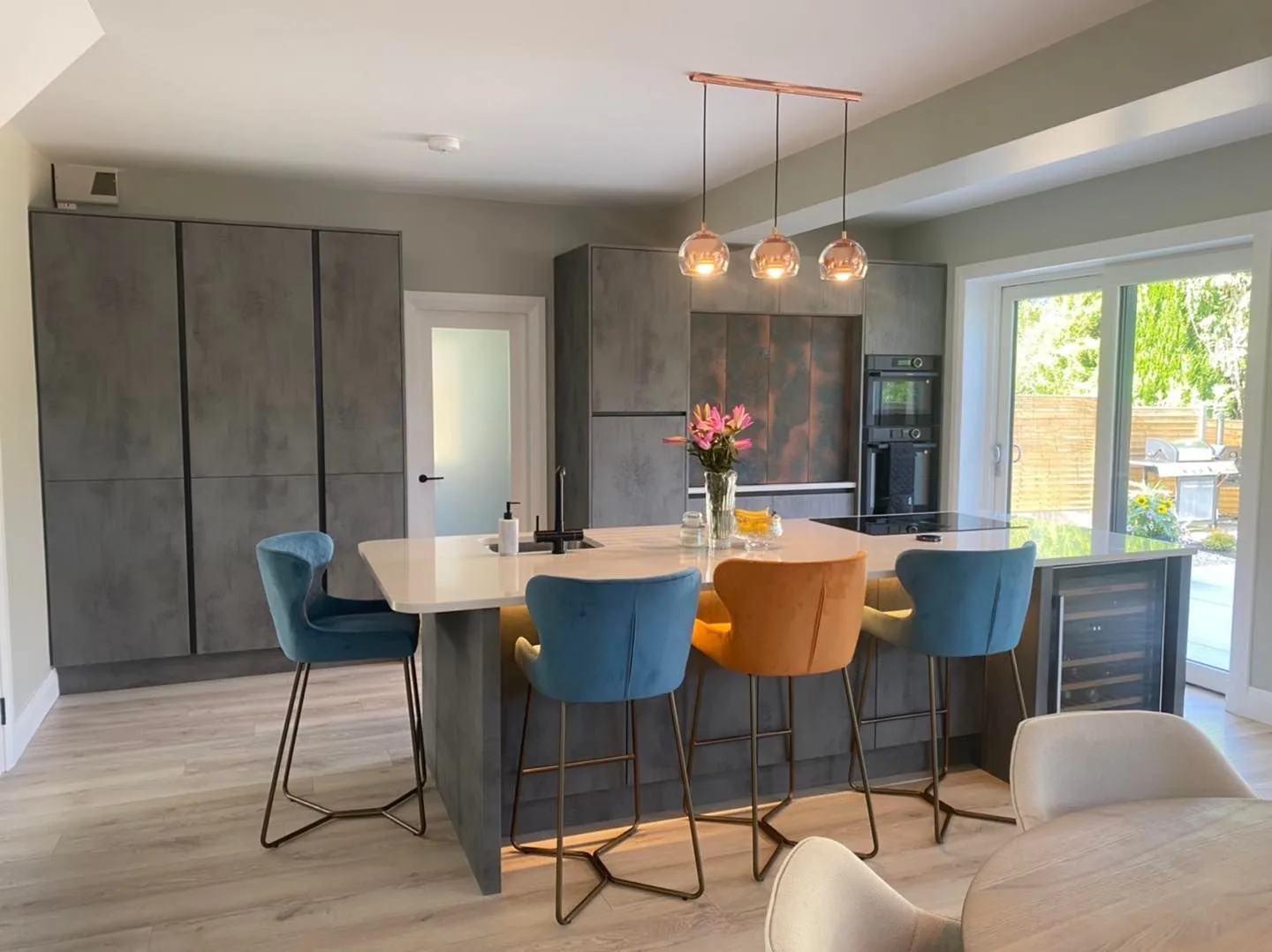 Concrete-effect kitchen with copper pendants and teal + mustard velvet stools